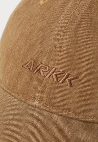 Casquette en velours côtelé beige avec un logo "ARKK" brodé en marron. Visière arrondie en tissu texturé avec des détails de couture visibles.