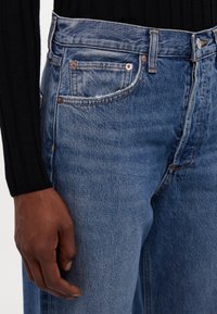 Modri džins hlače z rahlo zbledelim teksturo, opremljene s trdnimi bakrenimi dodatki in klasično zasnovo s petimi žepi. Vidna je roka na strani.