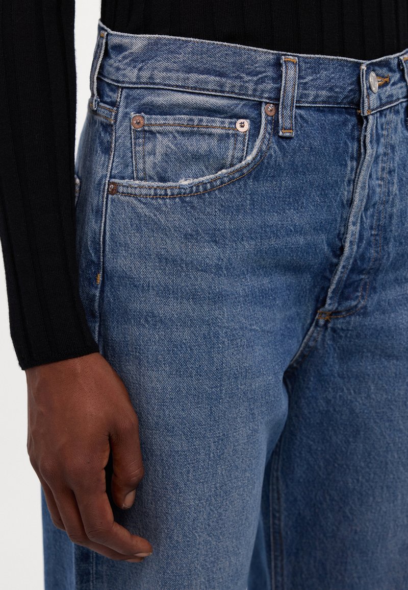 Blauwe spijkerbroeken van denim met een iets vervaagde textuur, voorzien van koperen hardware en een klassiek vijf-pocket ontwerp. Een hand is zichtbaar aan de zijkant.