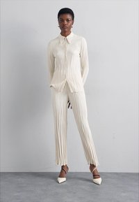 Cream-colored, pleated shirt and pants set with a collar. Pants have tapered hems. Model wears pointed shoes with ankle straps.