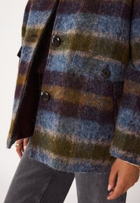 Cappotto oversize in misto lana con motivo a quadri blu, verde, marrone e viola, grandi bottoni neri e una finitura morbida e testurizzata.