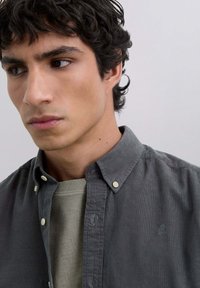 Camisa de botonadura gris de pana con botones blancos, llevada sobre un suéter de cuello redondo gris claro. Detalle sutil del logo en la camisa.