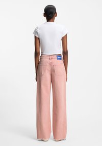 Vaaleanpunaiset väljälahkeiset farkut denimiä, joissa on klassinen viiden taskun malli ja sininen logoplässi takavyötäröllä.