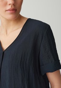 Tessuto blu navy, stropicciato, maniche corte con risvolti, scollatura a V e vestibilità comoda. La texture sembra morbida e leggera.
