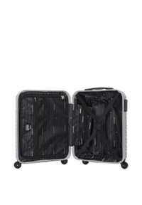 Hard-shell suitcase in silver, open to reveal black interior with linear pattern. Features cross straps and zippered compartments. Four wheels.
