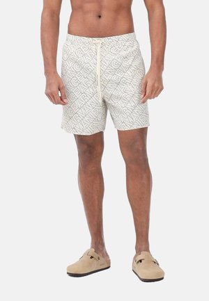 Uomo che indossa pantaloncini beige con coulisse a motivo e zoccoli beige senza lacci, in piedi contro uno sfondo chiaro e uniforme.