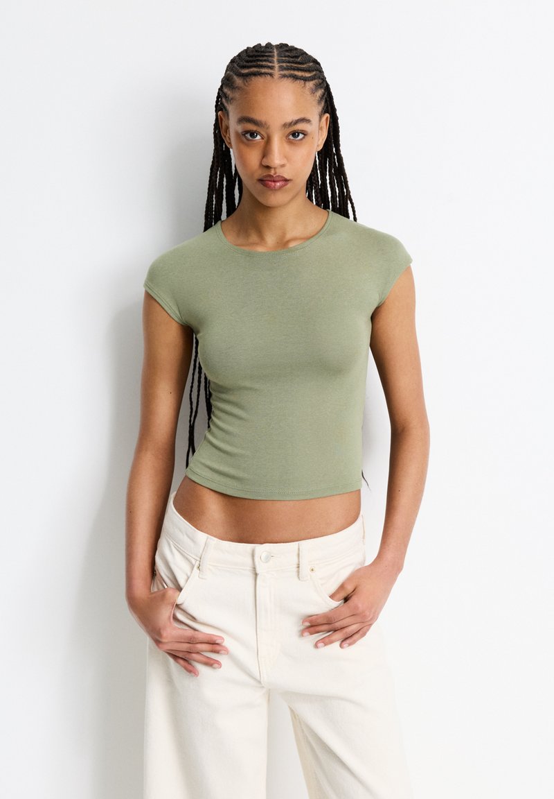 Kortärmad, figurnära grön topp i lätt jerseystoff. Har en rund halsringning och en kort design. Kombineras med högmidjade vita jeans.