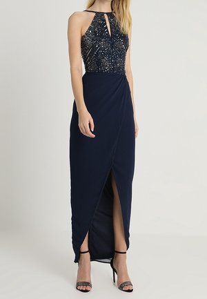 Granatowa sukienka na halterze z ozdobnym gorsetyem, przedni dekolt z wycięciem na kształt klucza, rozcięcie w kopertowej spódnicy, noszona z czarnymi sandałkami na wysokim obcasie z paskami.