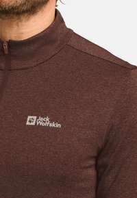 Brauner Mid-Zip-Fleece-Pullover mit hohem Kragen, der eine subtile Textur aufweist und ein weißes Jack Wolfskin-Logo im linken Brustbereich trägt.