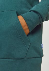 Felpa teal con tasca a marsupio, polsini e fondo ribattuti. Presenta un piccolo etichetta blu sul lato e un tessuto morbido e texturizzato.