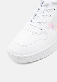 Sneaker bianco con tomaia sintetica liscia, punta rotonda, dettagli perforati e striscia laterale iridescente. Suola in gomma con texture e pattern antiscivolo.