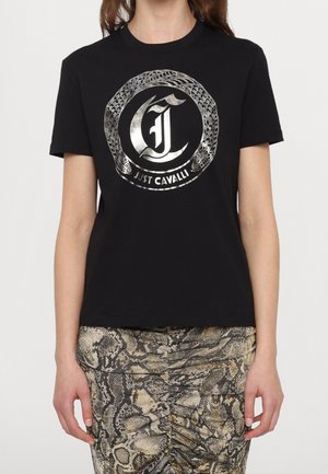 Nainen, joka käyttää mustaa t-paitaa, jossa on metallinen käärmekuvio ja "Just Cavalli" -teksti, yhdistettynä rypytettyyn käärmekuvioiseen hameeseen.