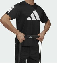 Camiseta negra de Adidas de manga corta con un gran diseño de logo blanco, combinada con pantalones cortos negros y sosteniendo una banda de resistencia.