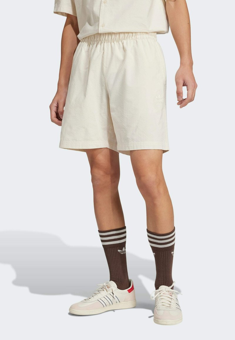 Light beige shorts with an elastic waistband, featuring a subtle texture and embroidered logo. Paired with brown striped socks and white sneakers.