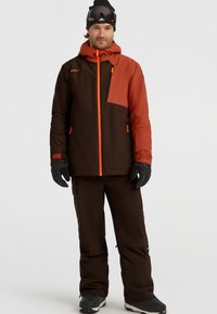 Skijack in bruin met oranje accenten, voorzien van een ritszak op de borst en een verstelbare capuchon; gecombineerd met een bijpassende bruine skibroek.