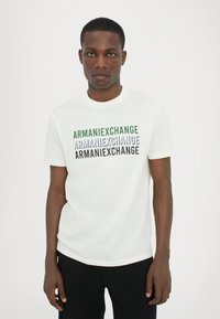 Balts īsajam piedurkņu T-kreklam ar apaļu kaklu, uz kura priekšpuses ir drukāts teksts "ARMANIEXCHANGE" zaļā, zilā un melnā krāsā.