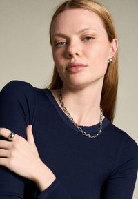 Dunkelblaues Ripp-Longsleeve mit Rundhalsausschnitt. Silberne Kettenhalskette mit miteinander verbundenen Gliedern. Das Modell trägt einen auffälligen Ring an ihrem Finger.