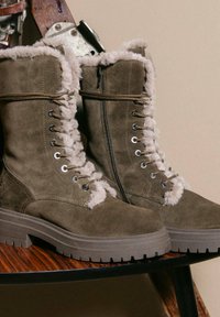 Brązowe zamszowe buty lace-up z futrzanym wykończeniem i grubą gumową podeszwą, umieszczone na drewnianym krześle na beżowym tle.