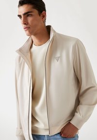 Jonge man in een beige zip-up jack over een crèmekleurige trui en blauwe jeans, die opzij kijkt met zijn hand in zijn zak, op een effen lichte achtergrond.