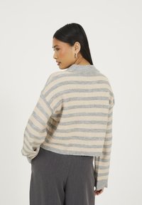 Kobieta z długimi czarnymi włosami, nosząca kolczyki w kształcie kół, beżowo-szaro paskowaną bluzkę i ciemnoszare spodnie, stoi bokiem z rękami w kieszeniach.