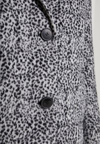 Szary i czarny płaszcz w kropki o miękkiej teksturze. Posiada dwa czarne guziki oraz kołnierz z wcięciem, podkreślający jego wzorzysty design.