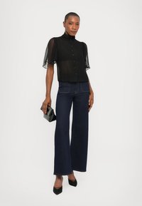 Black sheer blouse with puffed short sleeves, button closure, paired with dark blue flared jeans and black high-heeled shoes, holding a small black clutch.
