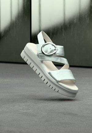 Zilveren sandaal met open teen, dikke zool, brede bandjes en een grote ronde gesp, hangend boven een grijs gestructureerd oppervlak.