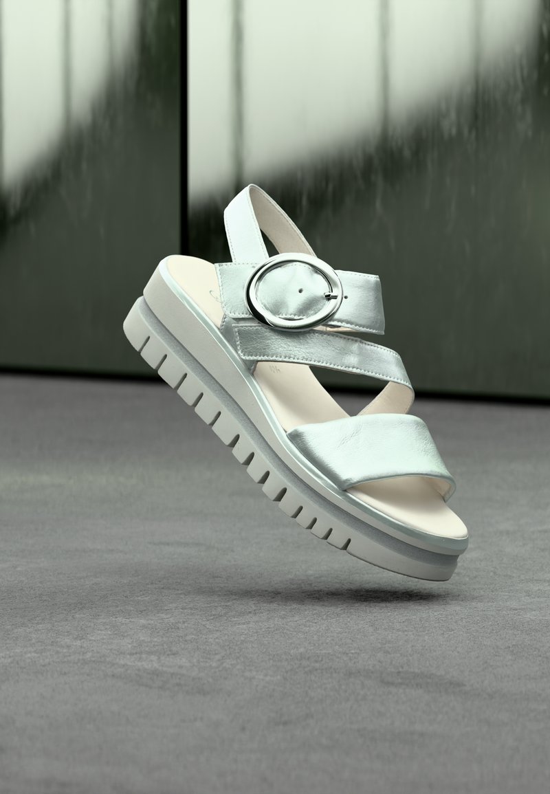 Sandale argentée à bout ouvert avec semelle épaisse, larges brides et grande boucle circulaire, suspendue au-dessus d'une surface texturée grise.