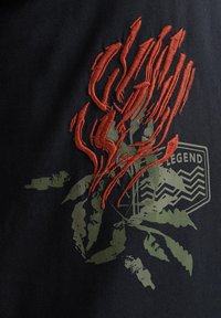 Černé tričko s texturovaným červeným vyšívaným květinovým vzorem a zeleným potiskem listí, se znakem obsahujícím slovo "LEGEND."