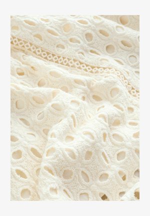 Tissu blanc brodé avec des motifs découpés, des détails texturés et une délicate bordure en dentelle, créant un design motif.