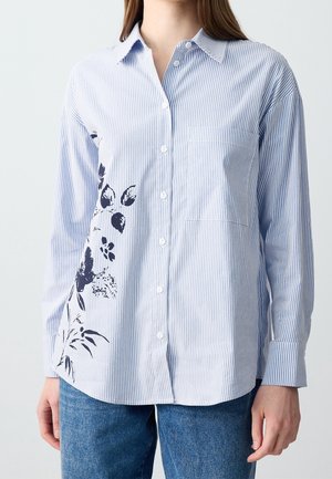 Femme portant une chemise boutonnée à rayures bleu clair et blanc avec un motif floral noir sur le côté gauche, associée à un jean bleu.