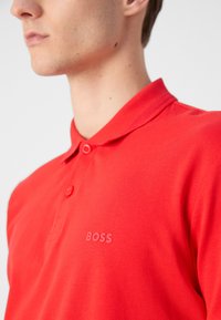Camisa polo roja hecha de tela texturizada, con un cierre de tres botones y un sutil logo en relieve en el lado izquierdo.