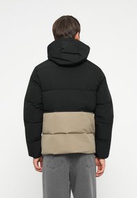 Padded jacket with a black upper section and beige bottom; features a hood and a boxy silhouette. The fabric appears smooth and lightweight.