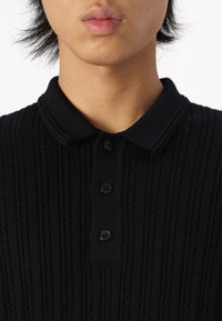 Polo de punto negro con canalé vertical, tres botones negros en el cuello y una tela texturizada. Sin patrones ni acentos visibles.