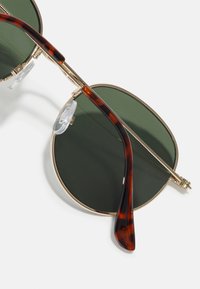 Lunettes de soleil rondes avec des verres vert foncé, monture en métal doré et branches à motif écaille de tortue. Équipées de coussinets de nez transparents pour plus de confort.