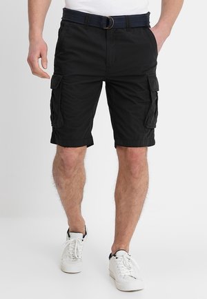 Uomo che indossa pantaloncini cargo neri con tasche laterali, una cintura blu scuro, scarpe da ginnastica bianche e una maglietta bianca, in piedi davanti a uno sfondo semplice.