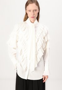 Blusa blanca con volantes y pliegues texturizados en el frente y las mangas, con cuello alto y puños fruncidos. El material parece suave y ligero.
