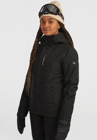 Veste étanche noire avec fermeture éclair à l'avant, capuche réglable et deux poches latérales. Portée avec des gants noirs et un bonnet beige.