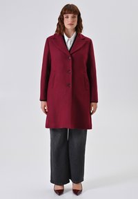 Vrouw in een bordeauxrode knopenjas over een wit shirt, zwarte wijde jeans en bordeauxrode puntige hakken, staand tegen een effen achtergrond.