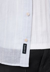 Vaaleansininen kevyt paita, jossa on pystysuorat raidat, napitettu beigenappi, ja jossa on AllSaints-logon etiketti helmassa.