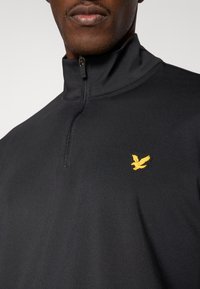 Pulover negru cu fermoar până la jumătate, realizat dintr-un material fin, având un mic logo galben cu pasăre pe piept și un guler drept.