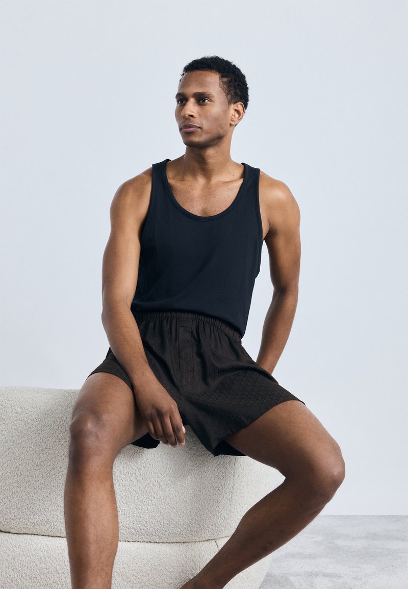 Must must tähnikate vasaku top ja pruunide mustriliste lühikeste pükste, istudes tekstuuriga heledat värvi diivanil, pehme halli tausta ees.