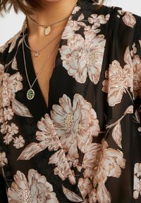 Czarna kwiatuszkowa kopertowa sukienka z dużymi jasnoróżowymi kwiatami z akcentami złota. Przezroczysty, lekki materiał z dekoltem w serek, w zestawie wielowarstwowe naszyjniki.