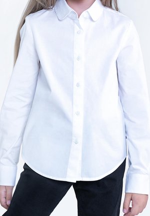 Niño con camisa blanca de manga larga abotonada y pantalones negros, de pie contra un fondo liso y claro.