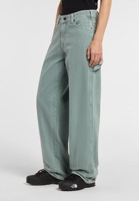 Ljusgröna jeans med vida ben och sliten textur, fem fickor och svart metallbeslag. Matchade med svarta texturerade skor.