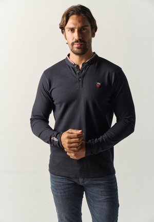 Hombre con cabello corto y castaño y barba, que lleva una camisa negra de manga larga y vaqueros azules, de pie frente a un fondo claro y liso.