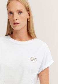 Weißes Baumwoll-T-Shirt mit umgeschlagenen Ärmeln, mit einem kleinen, braunen Drucktext, der "PASSIONE GODEMENTO COMUNITÀ" lautet. Glatte Textur.
