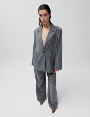 Sötét, hátrafésült hajjal rendelkező nő, aki túlságosan nagy, szürke tűszínű öltönyt és hegyes barna cipőket visel, egyszerű fehér háttér előtt.