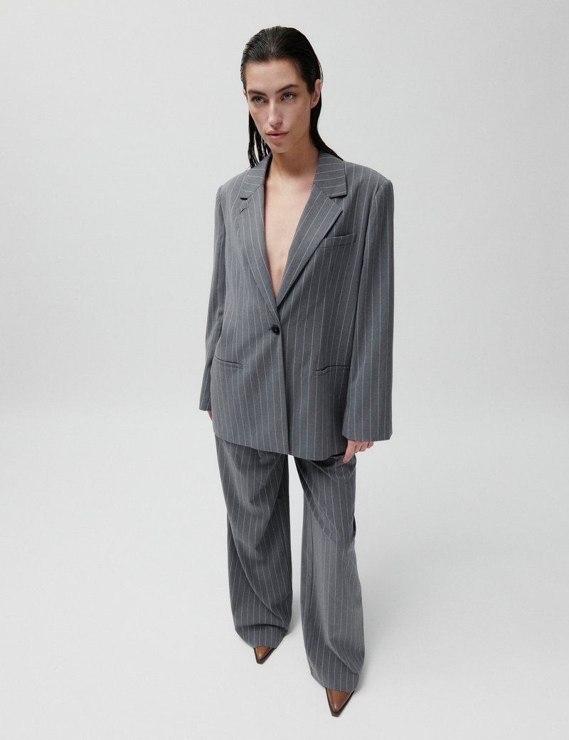 Γυναίκα με πίσω χτενισμένα σκουρόχρωμα μαλλιά, ντυμένη με υπερμεγέθη γκρι κοστούμι με ρίγες και μυτερά καφέ παπούτσια, σε απλό λευκό φόντο.