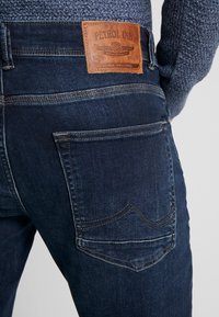 Jeans de mezclilla azul oscuro con una etiqueta de cuero que dice "Petrol Ind" en la parte posterior, con costuras de contraste y diseño estándar de bolsillo trasero.